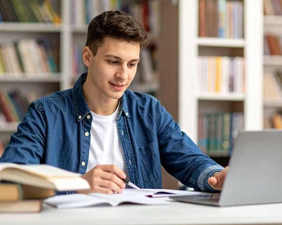 Estudiante con su portátil y sus libros en una biblioteca, preparando sus estudios